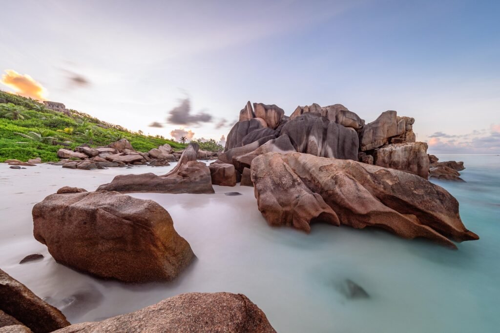 Scenic view of large granite boulders on a serene beach at sunset in Seychelles, with soft white sand and gentle waves lapping at the shore.